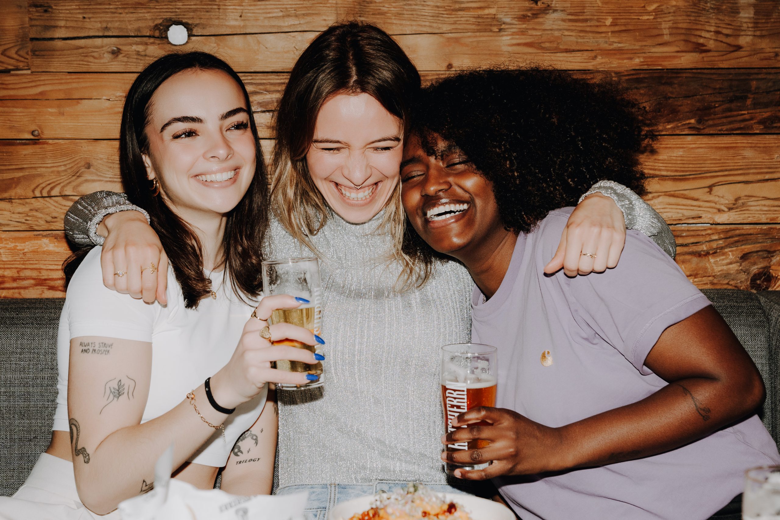 Drei junge Frauen sitzen lachend mit Bier in der Hand vor Nachos in einem Restaurant.